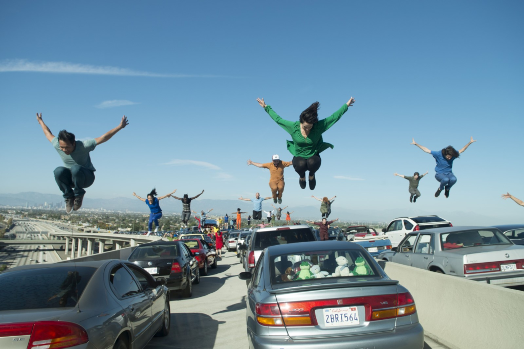 Cena de La La Land. Um grupo de pessoas está saltando em cima de carros que estão em uma ponte em Los Angeles.